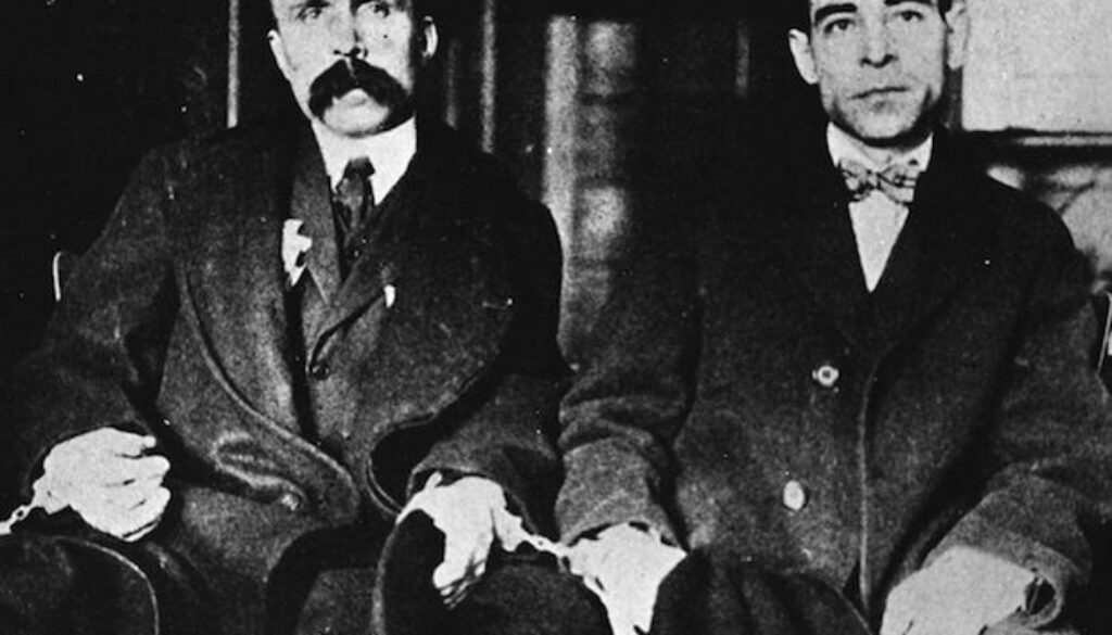 Sacco and Vanzetti, seated in handcuffs.